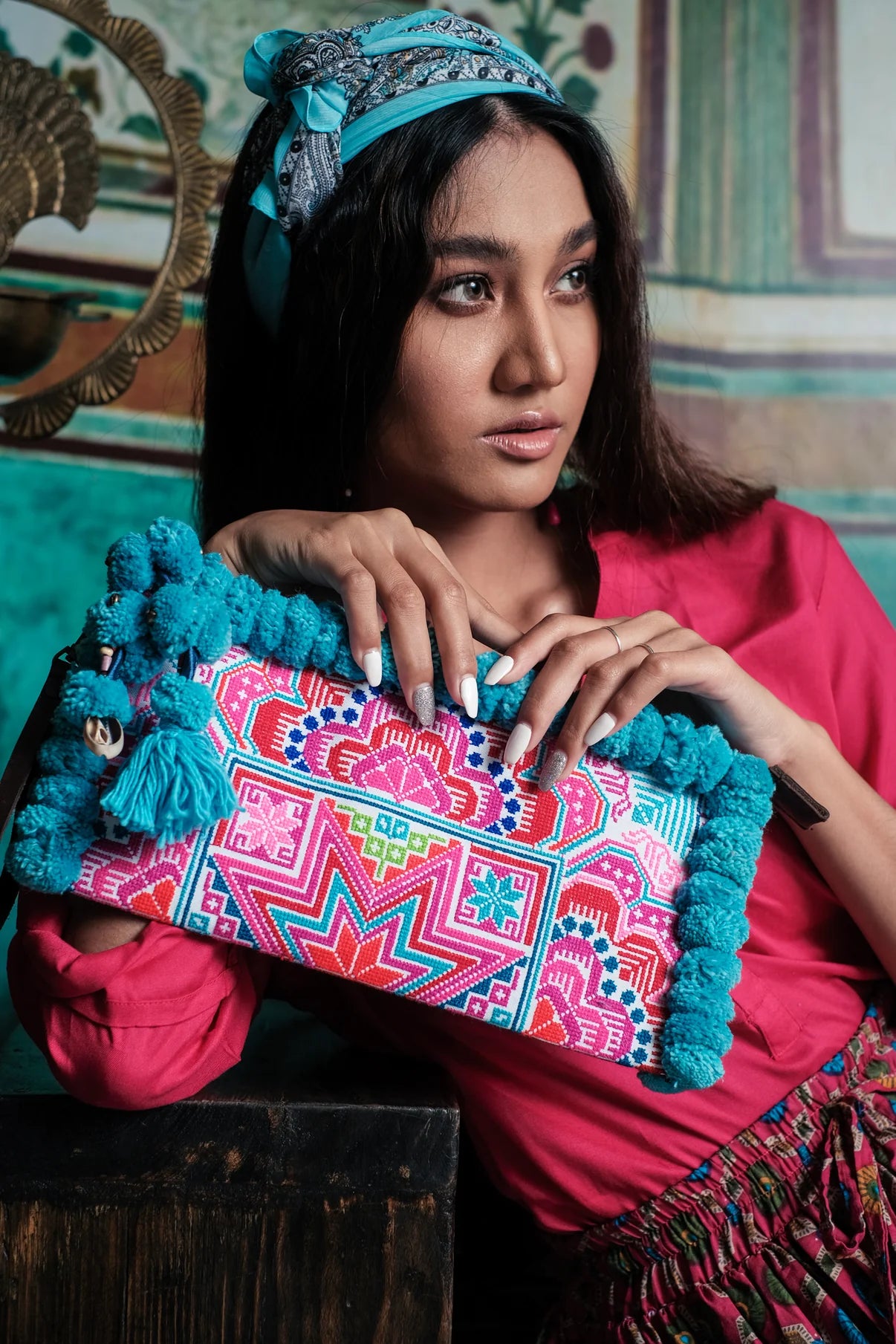 A woman poses elegantly with the Pom Pom Clutch Bag from Suzys Treasures, styled in bright colors with a turquoise headscarf. The embroidered clutch adds a burst of color and artisanal flair to her boho-chic outfit.