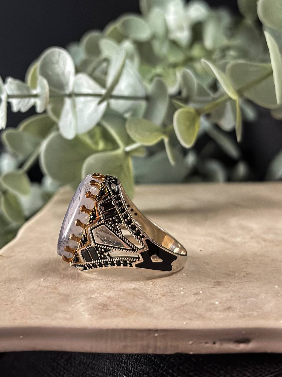 Suzys Treasures handcrafted Yemeni men’s ring featuring a Aqeeq (agate) stone set in an ornate sterling silver band with traditional motifs, displayed on a stone surface with a green leafy background.