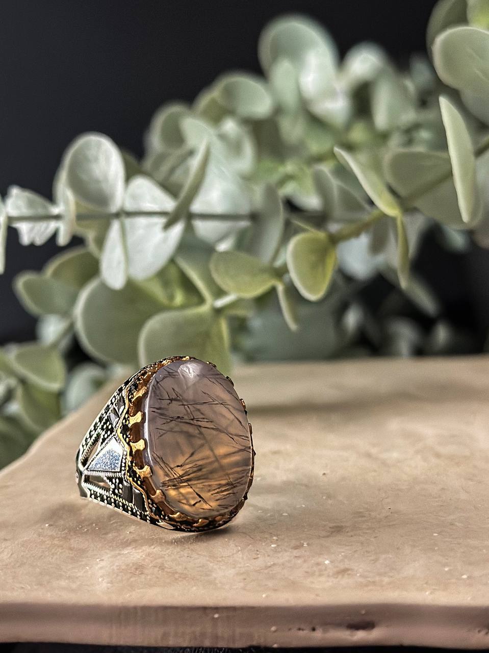 Suzys Treasures handcrafted Yemeni men’s ring featuring a Aqeeq (agate) stone set in an ornate sterling silver band with traditional motifs, displayed on a stone surface with a green leafy background.
