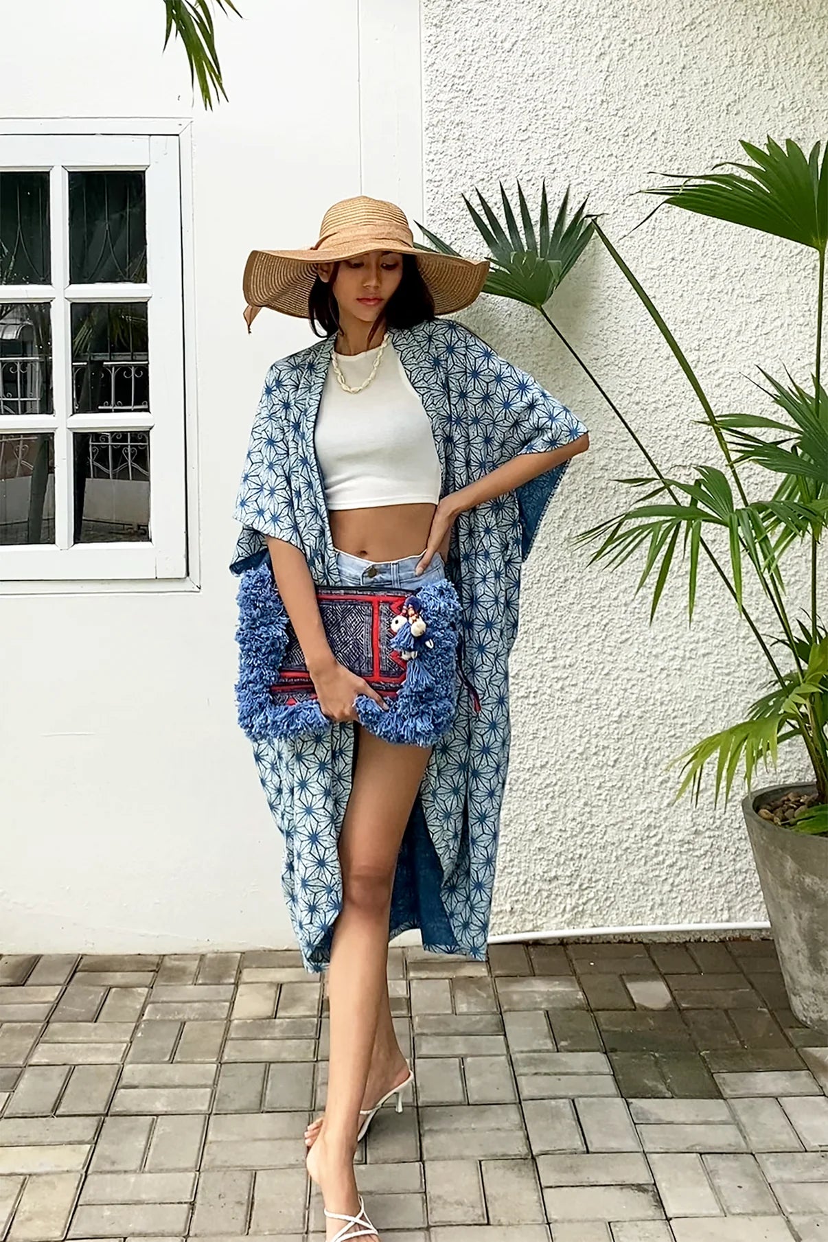 A woman poses in a breezy summer look wearing the Indigo Stars Kimono from Suzys Treasures, paired with a white crop top, denim shorts, and a straw hat. She carries a vibrant handmade clutch with blue fringe and tribal details, standing against a white wall and tropical greenery.