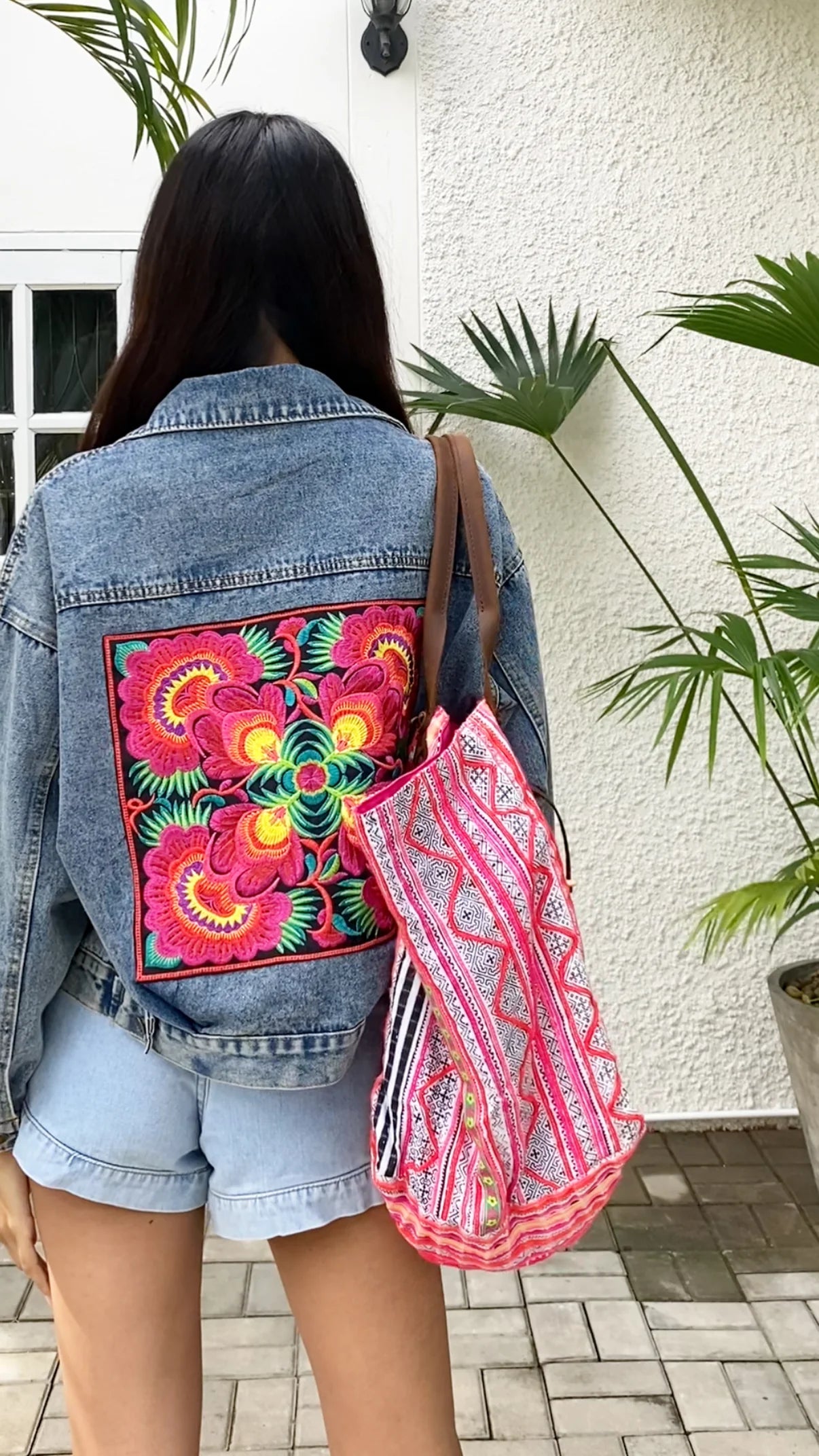 A woman shows off the Bohemian Denim Jacket with Hmong Embroidery from Suzys Treasures, featuring a bold floral panel in vibrant pink, yellow, and green on the back. Paired with casual shorts and a handwoven tribal sling bag, this look perfectly blends boho flair with traditional artisan craftsmanship.
