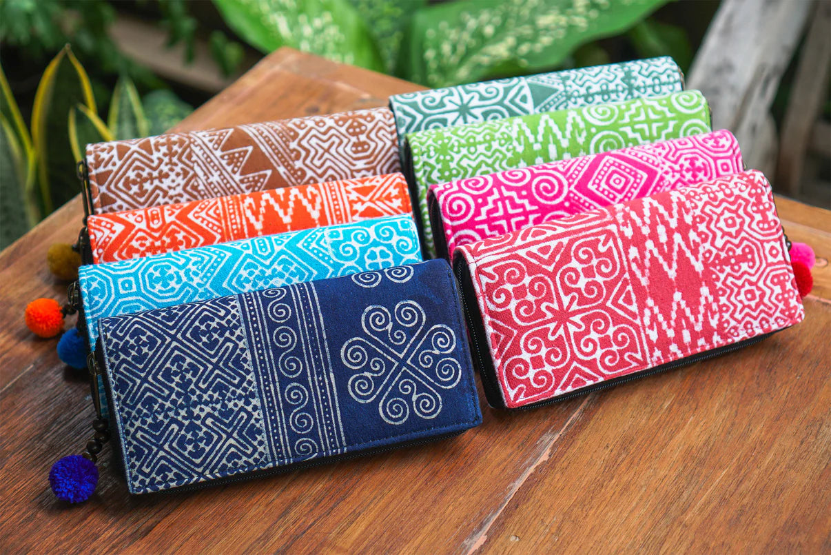 A colorful collection of handmade wallets from Suzys Treasures, each featuring bold tribal-inspired patterns in vibrant hues like navy, turquoise, red, pink, green, orange, and brown. Finished with playful pom-pom zipper pulls, these wallets beautifully blend tradition, function, and joyful design.