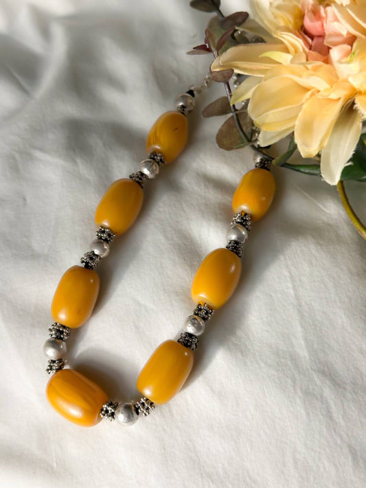 Suzys Treasures handcrafted Yemeni necklace with chunky honey-toned amber beads and ornate silver spacers, styled on a soft white fabric background with yellow flowers and foliage.
