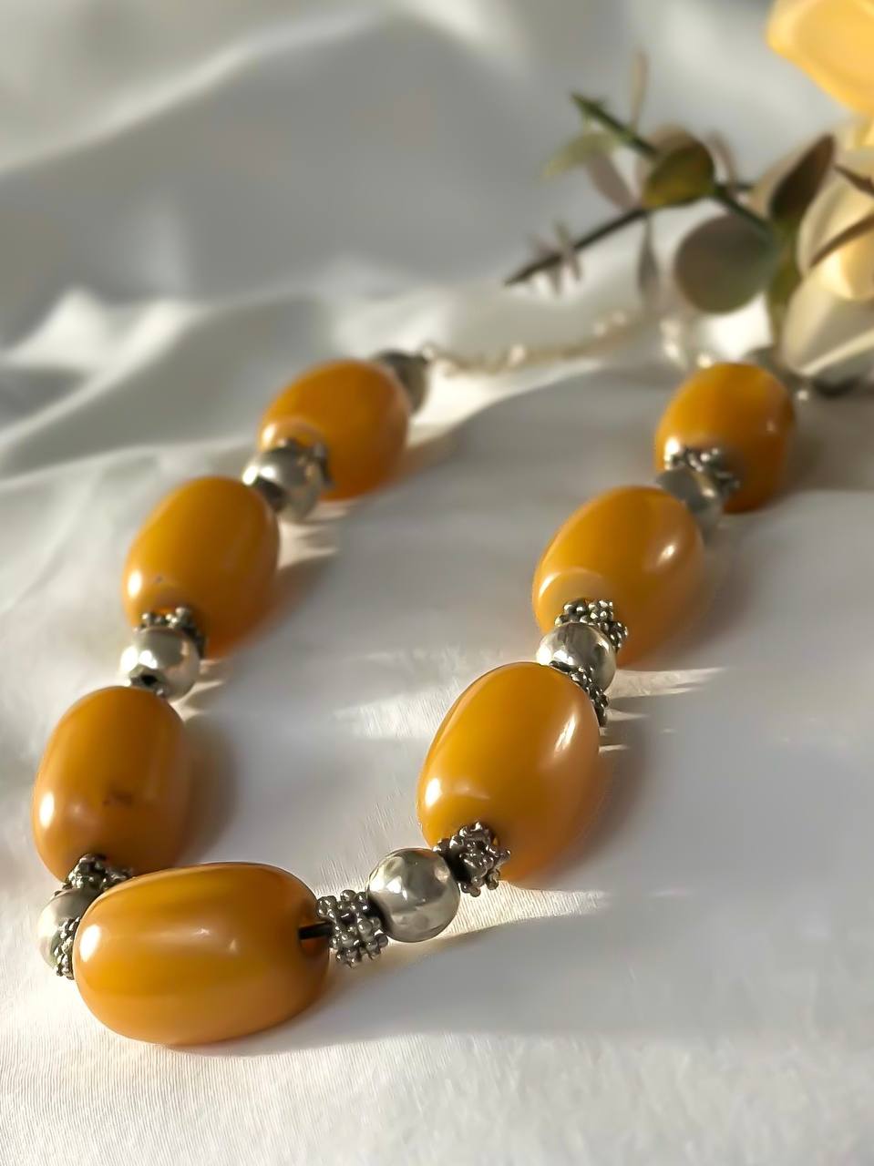 Suzys Treasures handcrafted Yemeni necklace with chunky honey-toned amber beads and ornate silver spacers, styled on a soft white fabric background with yellow flowers and foliage.
