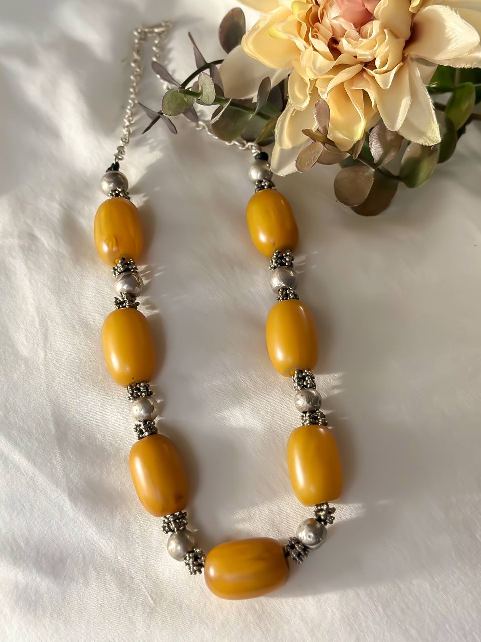 Suzys Treasures handcrafted Yemeni necklace with chunky honey-toned amber beads and ornate silver spacers, styled on a soft white fabric background with yellow flowers and foliage.