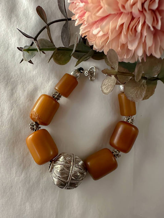 Suzys Treasures Yemeni Amber & Silver Talisman Bracelet featuring bold honey-toned resin beads and a handcrafted silver talisman charm, styled with soft pink blooms on a white background.