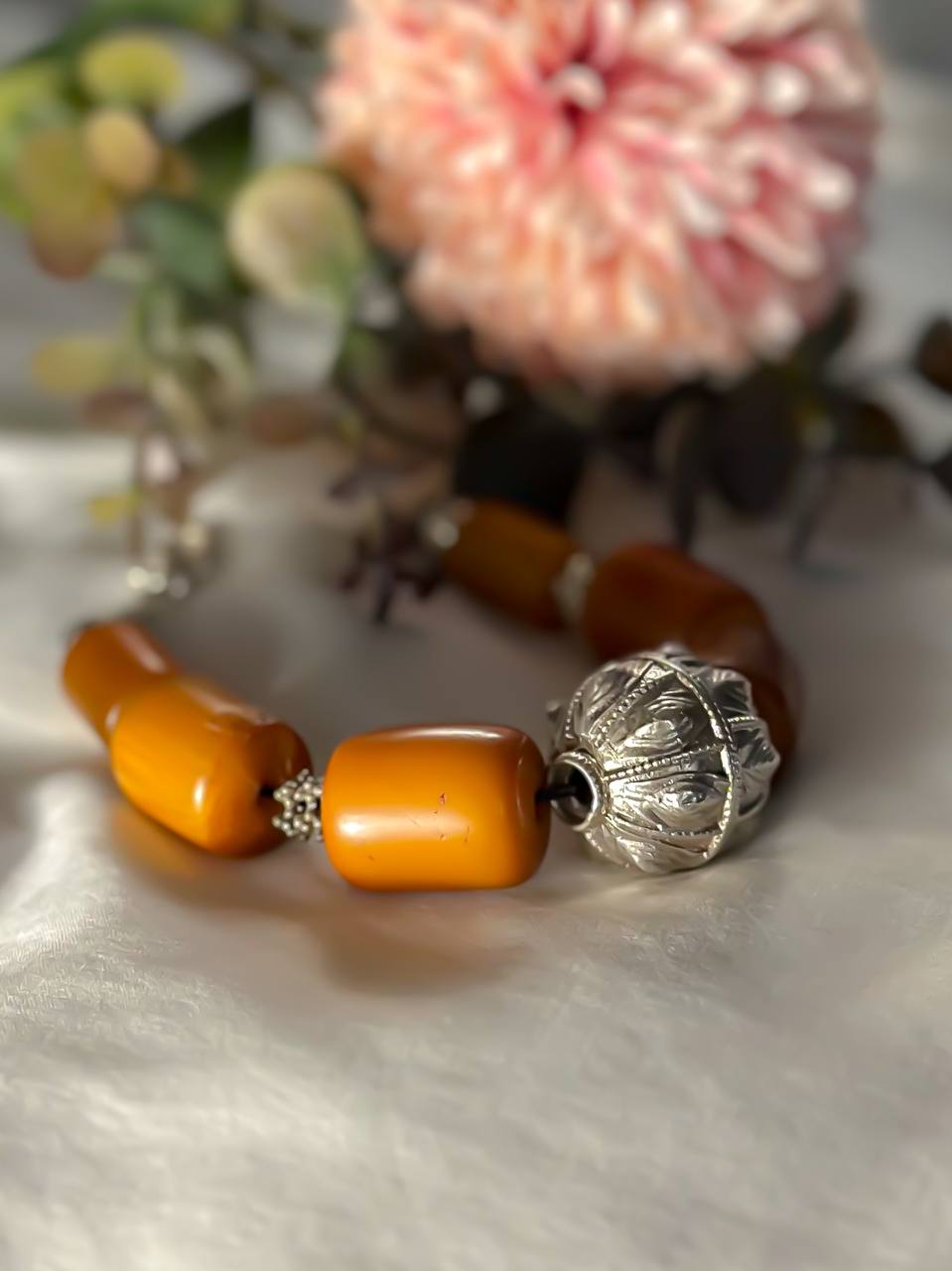 Suzys Treasures Yemeni Amber & Silver Talisman Bracelet featuring bold honey-toned resin beads and a handcrafted silver talisman charm, styled with soft pink blooms on a white background.