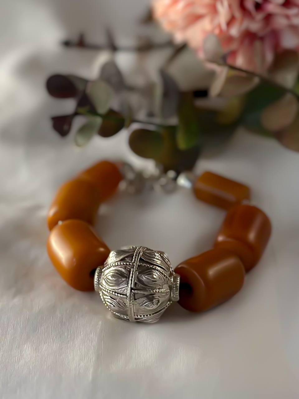 Suzys Treasures Yemeni Amber & Silver Talisman Bracelet featuring bold honey-toned resin beads and a handcrafted silver talisman charm, styled with soft pink blooms on a white background.