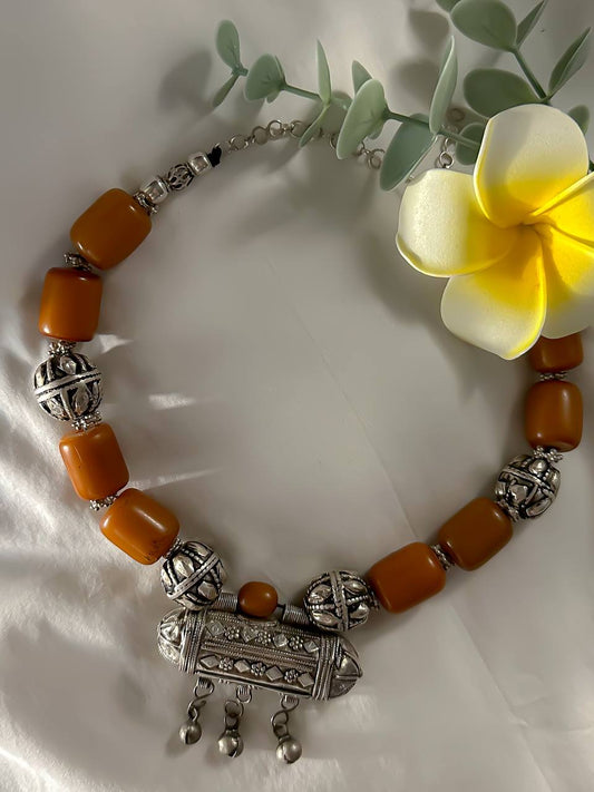 Suzys Treasures Yemeni Amber & Silver Hirz Amulet Necklace featuring chunky amber-hued beads and an ornate silver protective amulet pendant, styled with greenery and a yellow plumeria flower on white fabric.