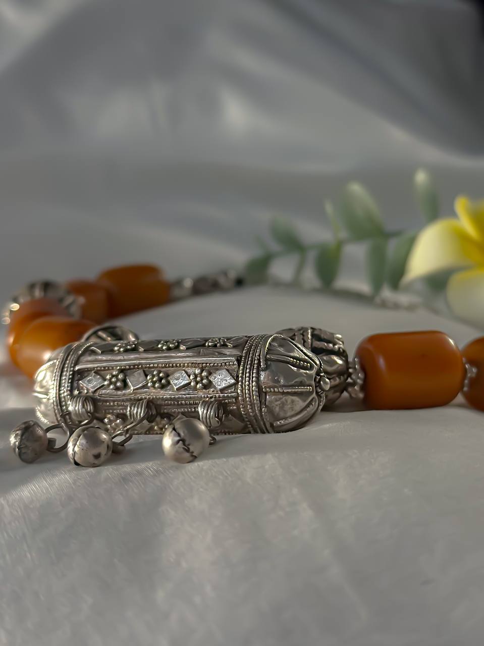Suzys Treasures Yemeni Amber & Silver Hirz Amulet Necklace featuring chunky amber-hued beads and an ornate silver protective amulet pendant, styled with greenery and a yellow plumeria flower on white fabric.