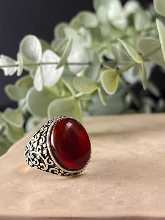 "Suzys Treasures handcrafted Yemeni men’s ring featuring a bold red Aqeeq (agate) stone set in an ornate sterling silver band with traditional motifs, displayed on a stone surface with a green leafy background.
