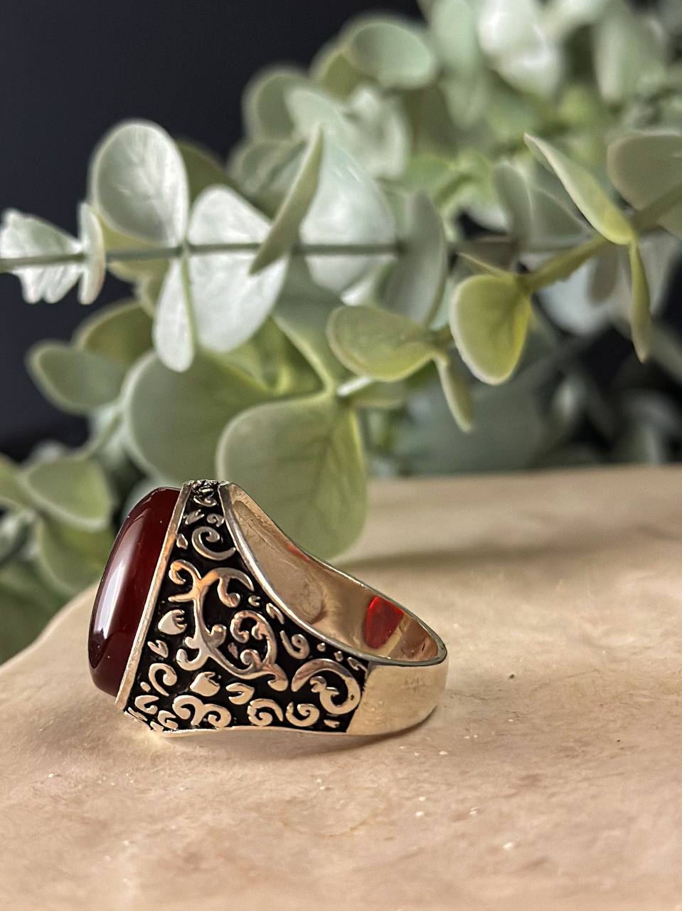 "Suzys Treasures handcrafted Yemeni men’s ring featuring a bold red Aqeeq (agate) stone set in an ornate sterling silver band with traditional motifs, displayed on a stone surface with a green leafy background.
