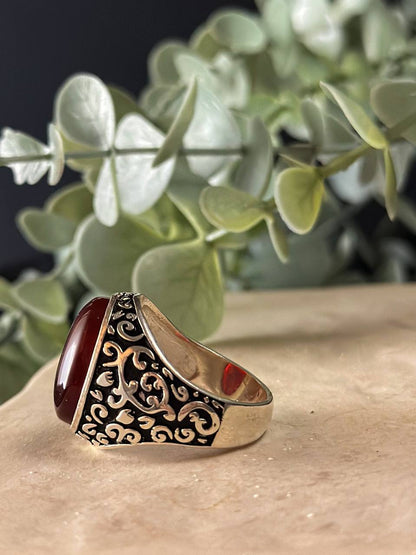 "Suzys Treasures handcrafted Yemeni men’s ring featuring a bold red Aqeeq (agate) stone set in an ornate sterling silver band with traditional motifs, displayed on a stone surface with a green leafy background.