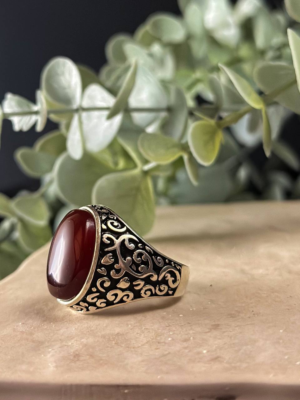 "Suzys Treasures handcrafted Yemeni men’s ring featuring a bold red Aqeeq (agate) stone set in an ornate sterling silver band with traditional motifs, displayed on a stone surface with a green leafy background.