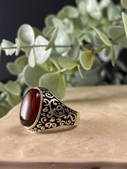 "Suzys Treasures handcrafted Yemeni men’s ring featuring a bold red Aqeeq (agate) stone set in an ornate sterling silver band with traditional motifs, displayed on a stone surface with a green leafy background.