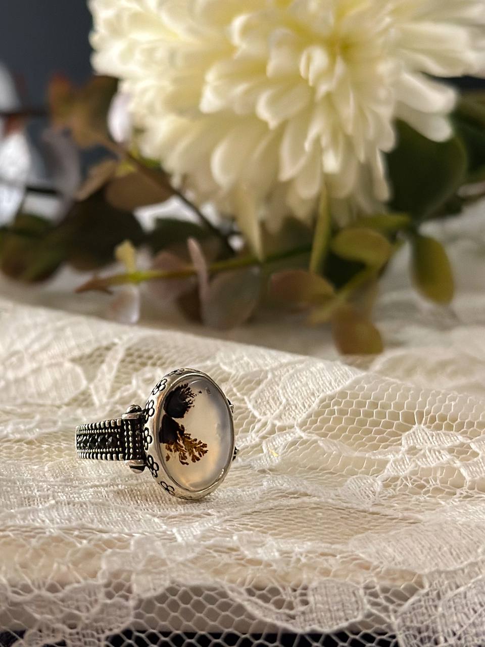 Suzys Treasures women's Yemeni dendritic agate ring with a detailed sterling silver band, displayed on delicate lace fabric with soft floral background.