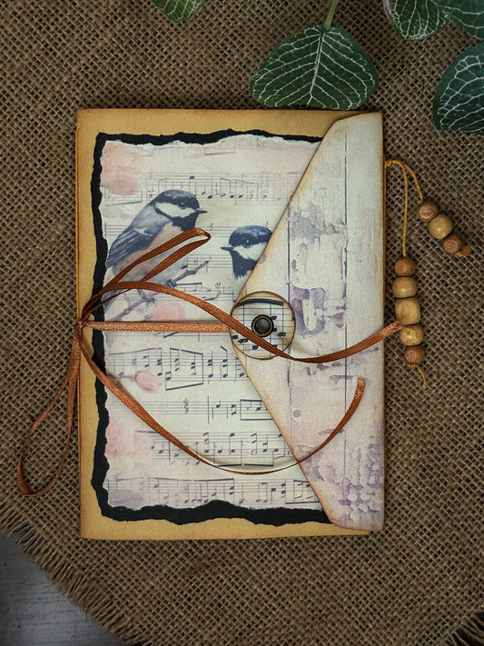 Suzys Treasures handcrafted brown vintage envelope journal featuring illustrations of birds and music notes, tied with leather cord and button closure, styled on burlap with wooden beads and green leaves.