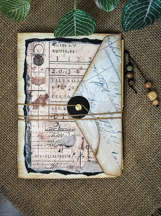 Suzys Treasures handcrafted brown vintage grunge mini envelope journal with distressed paper, twine closure, and aged handwriting design, styled on a burlap surface with decorative beads and leaves.