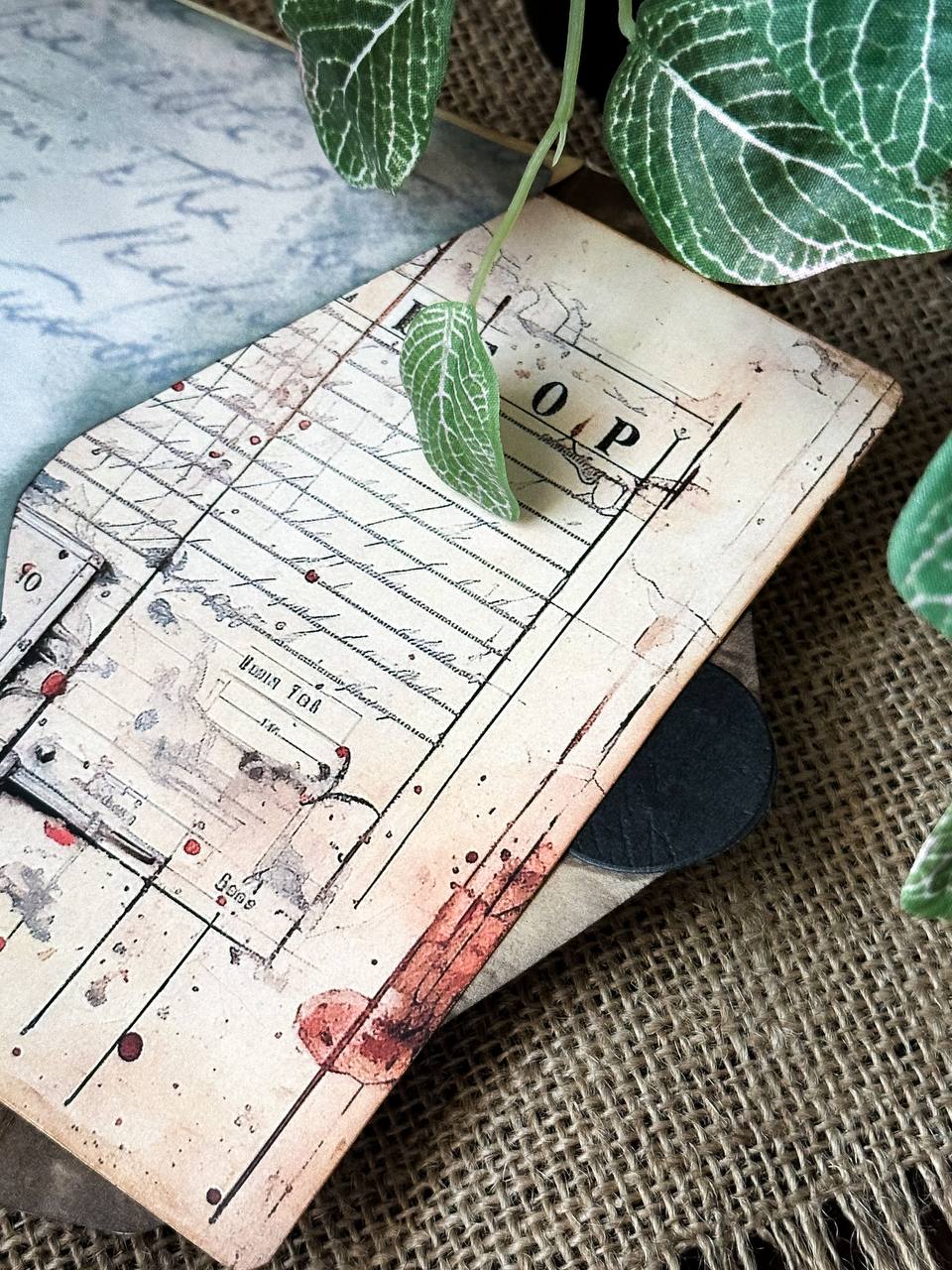 Detail shot of Suzys Treasures vintage grunge mini envelope journal, showing layered aged paper with French text and handwritten script, styled on burlap with green leaves.