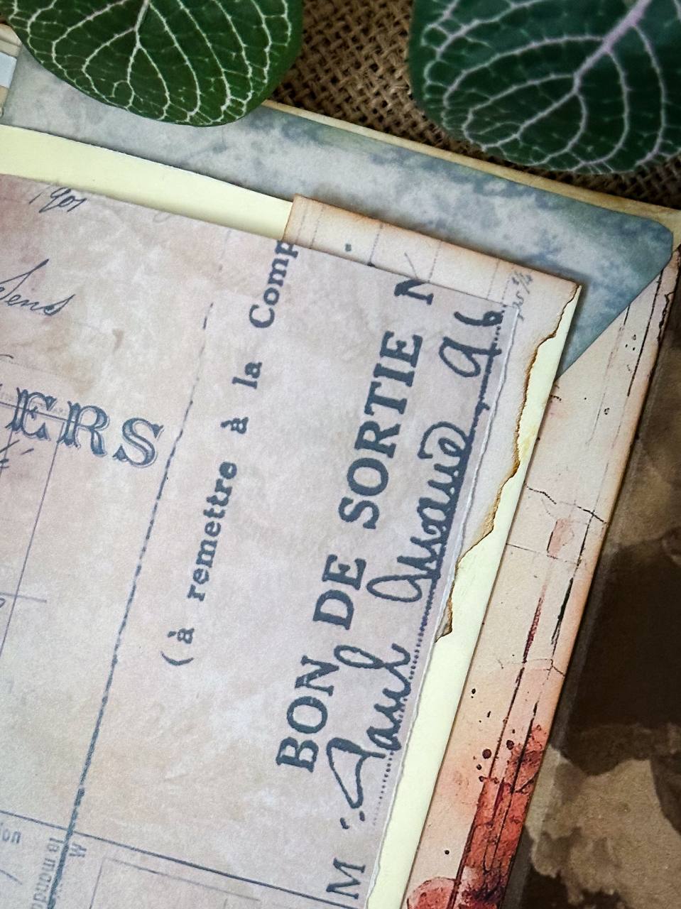Detail shot of Suzys Treasures vintage grunge mini envelope journal, showing layered aged paper with French text and handwritten script, styled on burlap with green leaves.