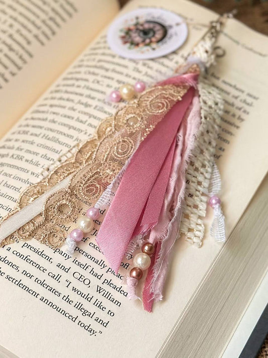 Handmade Boho tassel paperclip bookmark from Suzys Treasures, featuring pink and gold lace ribbons, pearl bead accents, and crochet trim, displayed on an open book – a charming and versatile accessory for journals, planners, or novels.