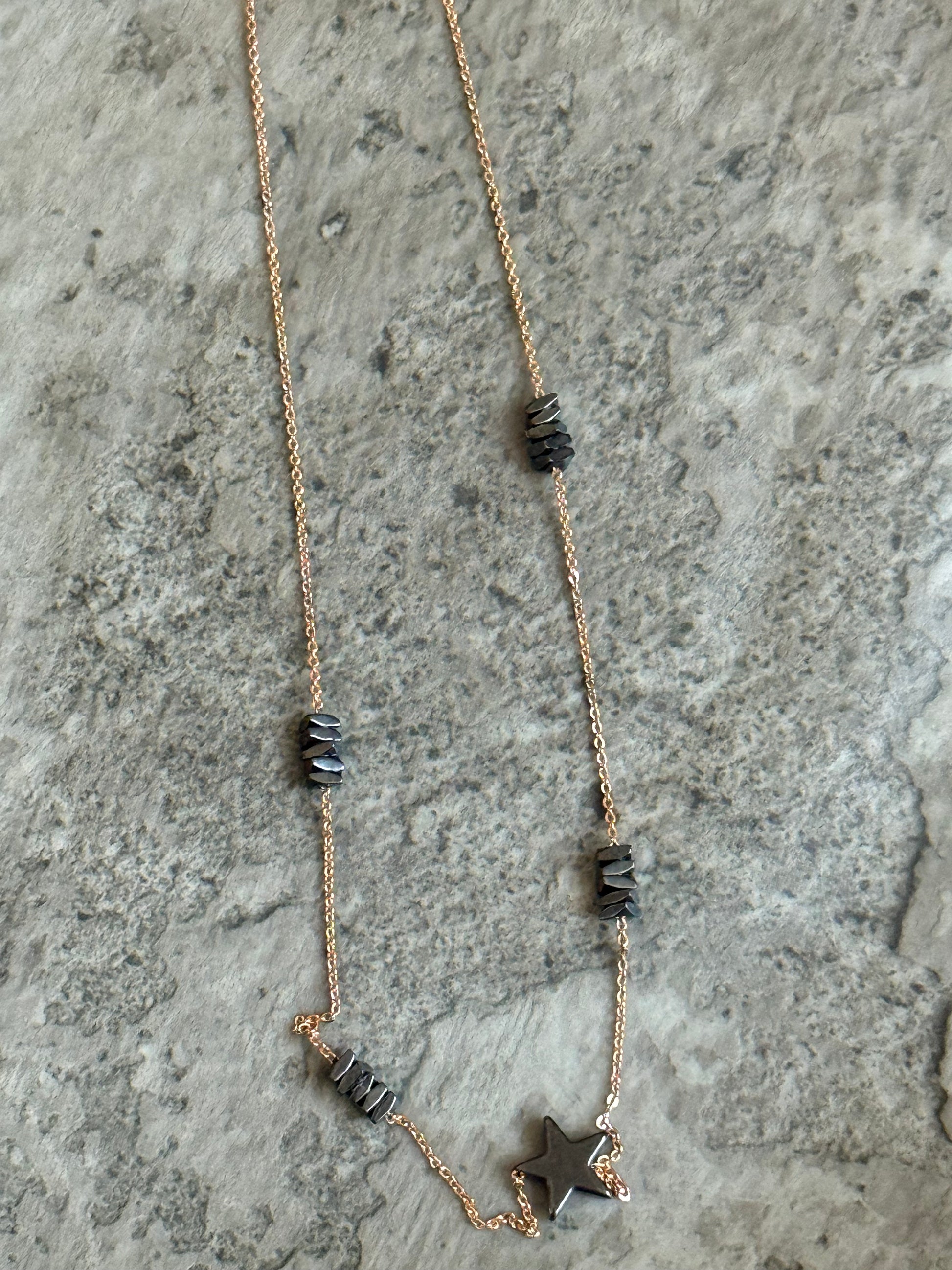Suzys Treasures handcrafted short necklace featuring stacked grey hematite beads and a hematite star charm on a silver embossed, warm-toned 3-micron plated chain, displayed on a textured stone background.