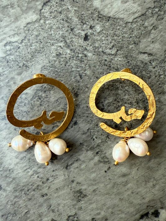 Gold-plated hammered earrings featuring the Arabic word “Hob” (حب) with dangling freshwater pearls, handcrafted and ethically made for Suzys Treasures, displayed on a textured stone background.