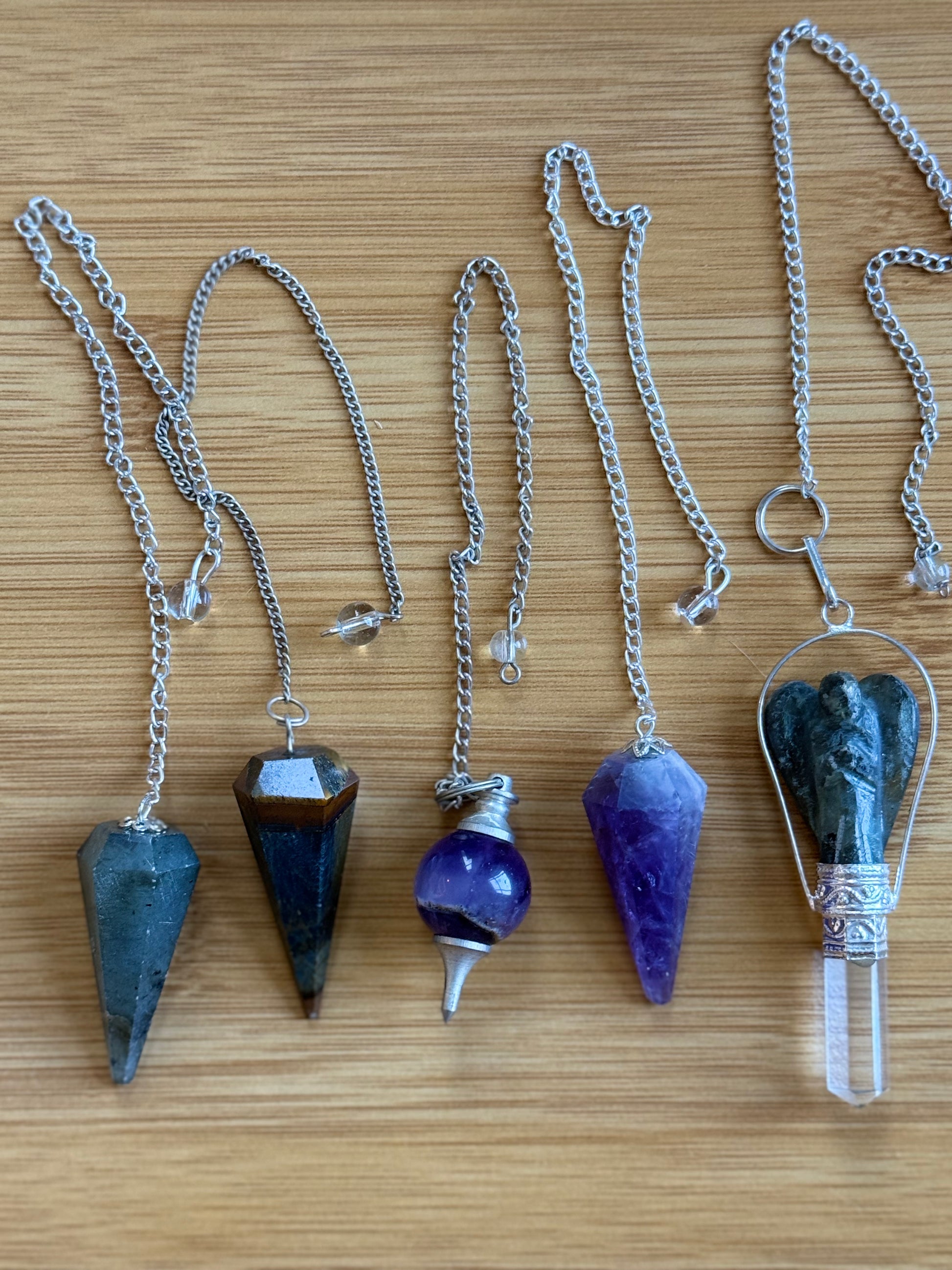 A set of five crystal pendulums laid out on a wooden background, including Labradorite, Tiger's Eye, Amethyst round, Amethyst long, and a Labradorite angel pendulum – Suzys Treasures.