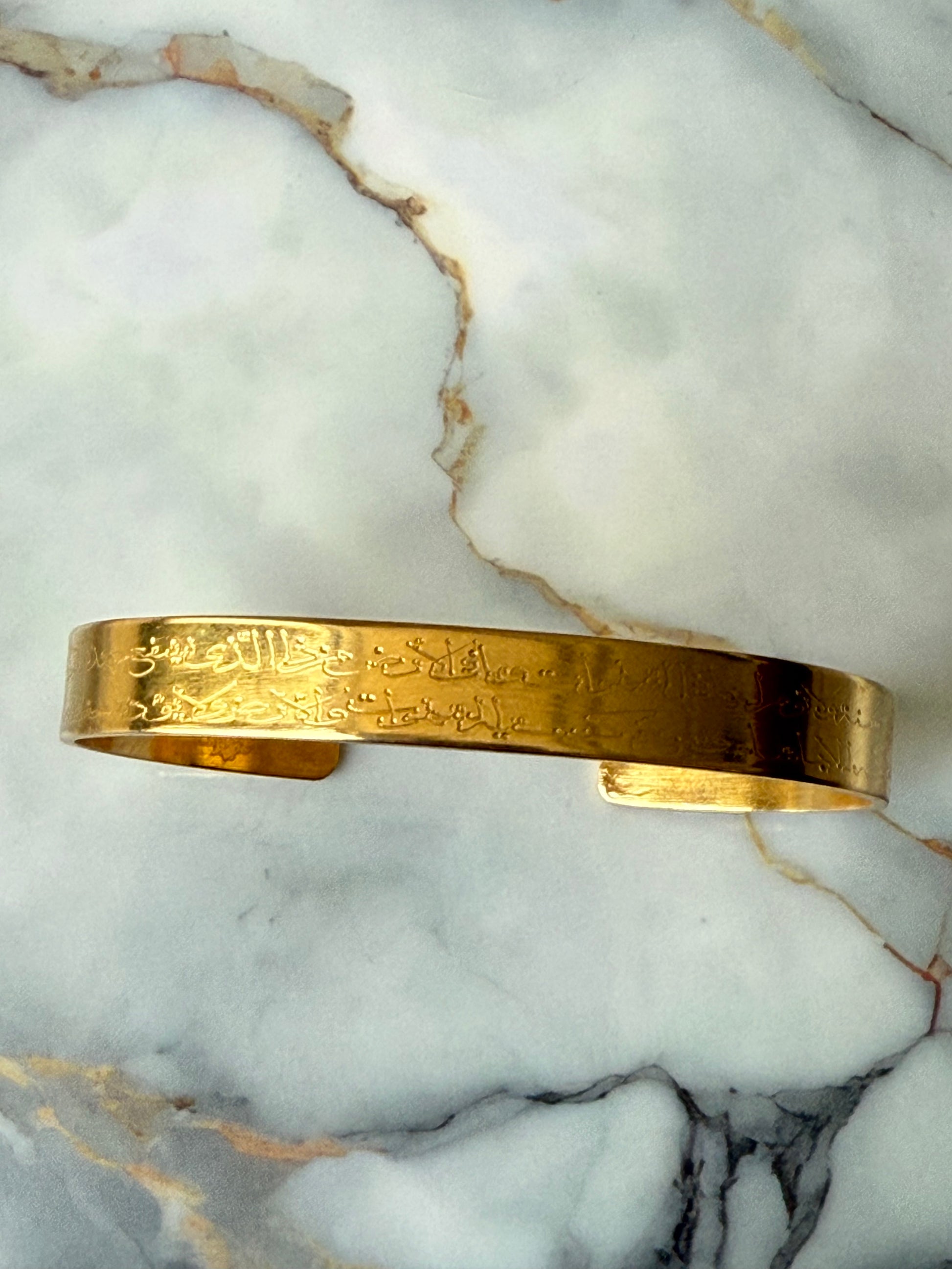 Thin gold-plated bangle engraved with Ayat Al-Kursi in Arabic script, a spiritual piece handmade for Suzy’s Treasures, photographed on a marble surface.
