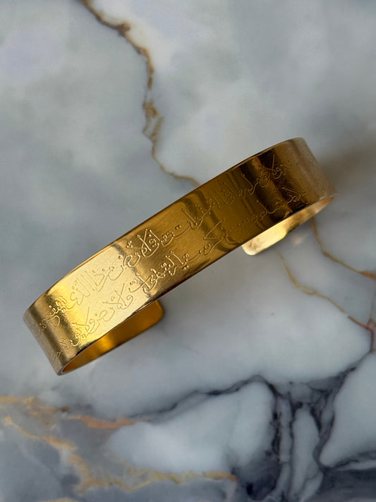 Wide gold-plated cuff bracelet with Ayat Al-Kursi engraved in elegant Arabic script, ethically crafted for Suzy’s Treasures, displayed on a marble background.