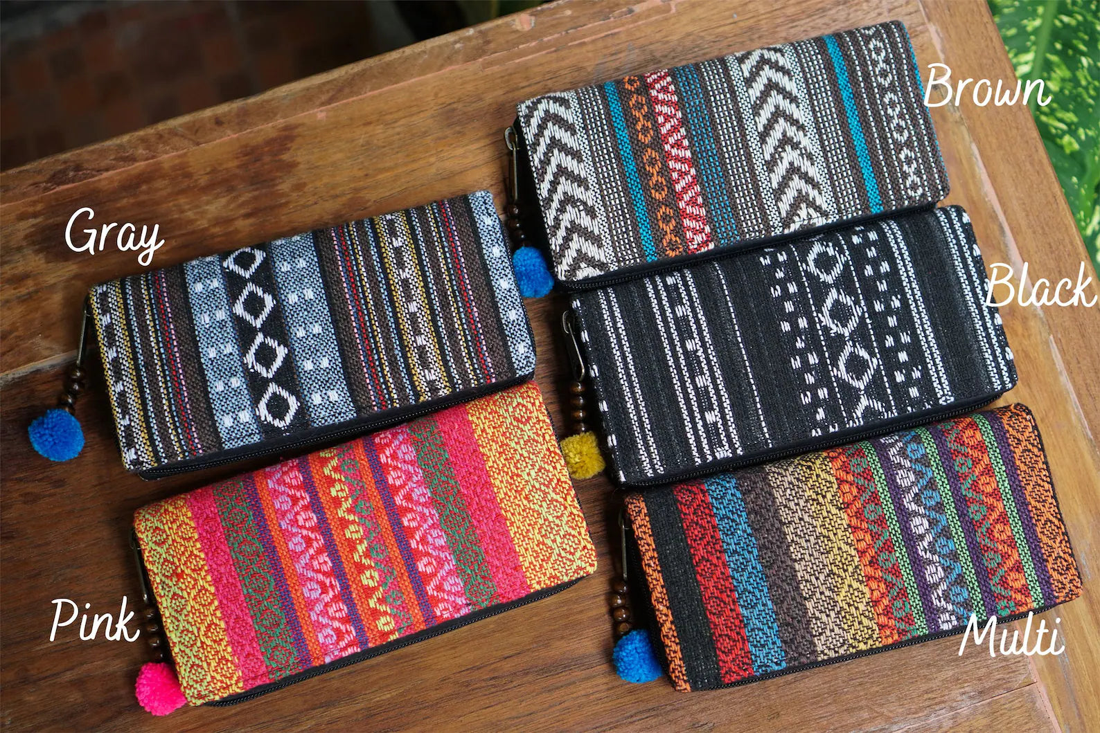 A colorful collection of handwoven wallets from Suzys Treasures, each with unique tribal-inspired patterns and playful pom-pom zippers. Available in Gray, Pink, Brown, Black, and Multi — perfect for adding charm to your everyday essentials.